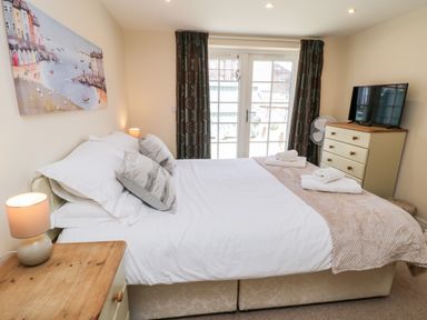 A bedroom with a bed with white linens and two pillows a wooden bedside table with a lamp a dresser with a television and a fan in front of glass doors with curtains at Razorbill Cottage Whitby