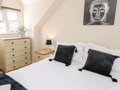 A bedroom with a bed white bedding and black pillows a wooden chest of drawers with a fan and a bedside table with a lamp and a painting on the wall at Razorbill Cottage Whitby