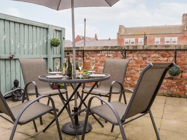 An outdoor patio with a round glass table and four chairs under an umbrella with a brick wall and green fence in the background at Razorbill Cottage in Whitby