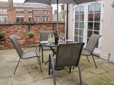 An outdoor patio area with a round glass table set with drinks and plates surrounded by four chairs and a large umbrella at Razorbill Cottage in Whitby