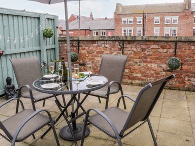A patio area with a round glass table set with glasses and plates surrounded by four chairs at Razorbill Cottage in Whitby