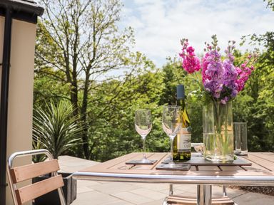 A patio table with a bottle of white wine two empty wine glasses and a vase of purple flowers surrounded by trees at Lands Brow Lodge in Wigton
