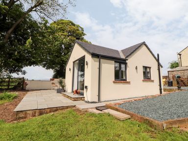 A small house with cream walls and dark roof tiles surrounded by grass and a paved area at Lands Brow Lodge in Wigton