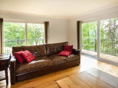 A living room with a brown leather sofa with red cushions and large glass sliding doors overlooking trees at Treetops in Govilon near Abergavenny