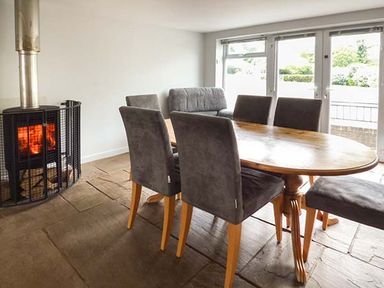 A dining area with a wooden table and six chairs near a fireplace with a fire and large windows at Treetops in Govilon near Abergavenny