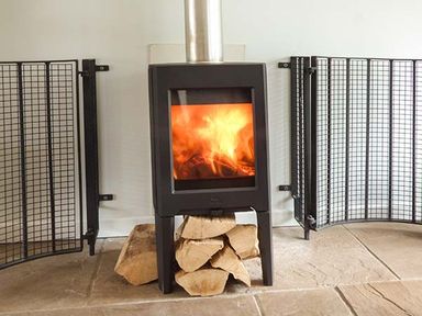 A burning wood stove with a black metal safety gate and stacked firewood underneath at Treetops in Govilon near Abergavenny