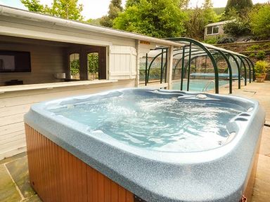 An outdoor hot tub with bubbling water next to a covered swimming pool and a white wooden building at Treetops in Govilon near Abergavenny