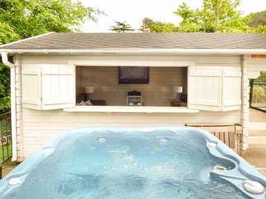 An outdoor hot tub in front of a white wooden cabin with open window shutters showing a room with lamps and a television at Treetops in Govilon near Abergavenny