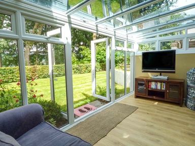 A conservatory with glass walls and roof a blue armchair a wooden TV stand with a flat screen TV and a beige rug on the floor at Treetops in Govilon near Abergavenny