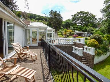 A patio with wooden lounge chairs and a black metal railing overlooking a garden with trees and bushes at Treetops Govilon near Abergavenny