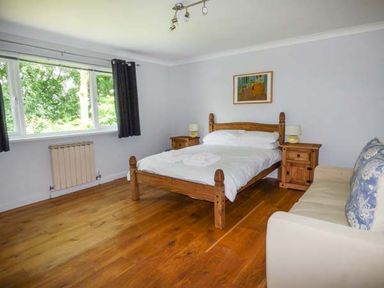 A bedroom with a wooden bed, two bedside tables with lamps, a couch, wooden floor, and window with dark curtains at Treetops Govilon near Abergavenny