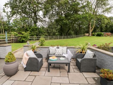 A patio with outdoor furniture consisting of two chairs and a sofa with cushions and throws around a table with drinks surrounded by plants and grass at Pondfield Gate in Cwmgors