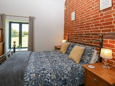 A bedroom with a bed beside nightstands and lamps by a window and a brick wall at The Long Barn in Morton on the Hill near Lenwade