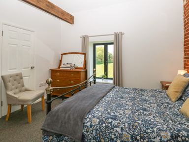A bedroom with a floral bedspread a wooden dresser a gray chair and a window with curtains at The Long Barn in Morton on the Hill near Lenwade