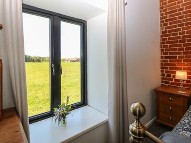 A bedroom window with cream curtains and a vase of flowers on the sill a brass bedpost and a wooden bedside table with a lamp at The Long Barn in Morton on the Hill near Lenwade