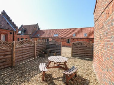 An outdoor gravel area with a wooden round picnic table and benches surrounded by wooden fencing at The Long Barn in Morton on the Hill near Lenwade