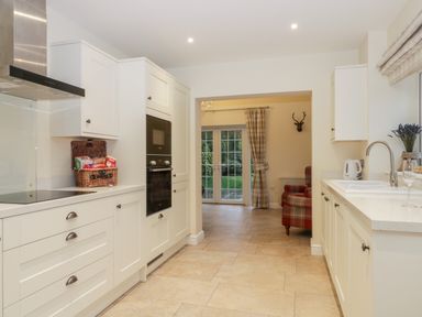 A kitchen with white cabinets an oven a wicker basket and a sink with a window showing a plaid armchair and glass doors at Lavender Cottage in Market Weighton
