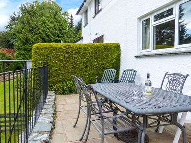 An outdoor patio with metal table and chairs a bottle and glasses on the table and a hedge near the house at Thompson Cottage in Ambleside