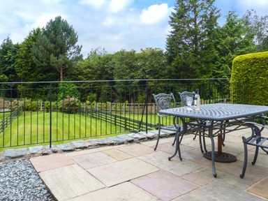 An outdoor patio with a metal table and chairs overlooking a grassy yard with trees at Thompson Cottage in Ambleside