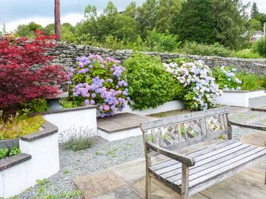 An outdoor garden area with a wooden bench stone wall and flowering shrubs at Thompson Cottage Ambleside