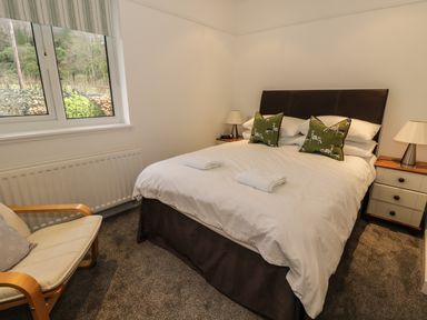A bedroom with a double bed with white bedding and green pillows with deer prints two bedside tables with lamps and a wooden armchair near a window at Thompson Cottage in Ambleside