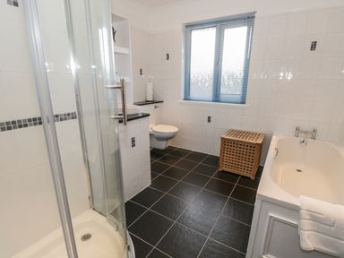 A bathroom with a shower, a bathtub, a wall mounted toilet, a window with blinds, and a wooden laundry basket at Thompson Cottage in Ambleside