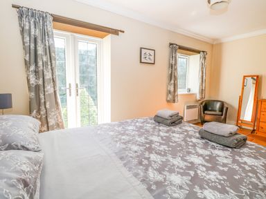 A bedroom with a bed covered in floral bedding two folded towels on the bed a window with curtains a leather chair a mirror and a wooden dresser at The Meadows Cottage in Billingford near Bawdeswell