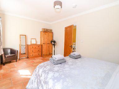 A bedroom with a bed covered in floral bedding folded towels on the bed a wooden wardrobe chest of drawers a standing mirror a brown armchair and a door at The Meadows Cottage in Billingford near Bawdeswell