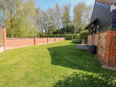 A backyard with green grass a brick and flint house a brick wall and trees at The Meadows Cottage in Billingford near Bawdeswell