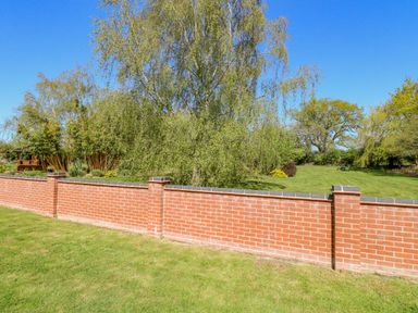 A red brick wall with grass and trees behind it at The Meadows Cottage in Billingford near Bawdeswell
