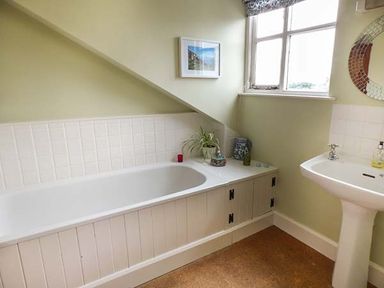 A bathroom with a bathtub under a slanted ceiling a pedestal sink a window and a round mirror at Gatehouse in Painscastle
