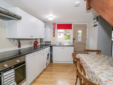 A kitchen with white cabinets black countertops a washing machine a dining table with a floral tablecloth and wooden chairs at The Coach House in Bitton