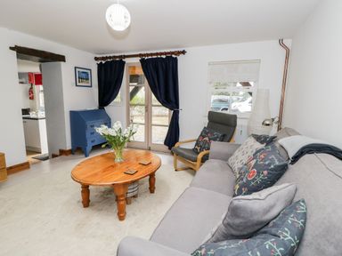 A living room with a gray sofa with cushions a wooden coffee table with flowers a gray armchair a blue chest of drawers and dark curtains at The Coach House in Bitton