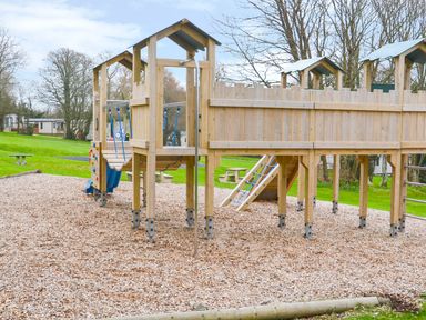 A wooden playground structure with slides climbing elements and wood chip flooring in a grassy outdoor area at High Tide in Rock
