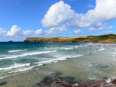 A beach with many people surfing and swimming in the water and a grassy coastal hill in the background at High Tide in Rock