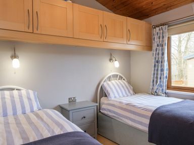 A bedroom with two single beds featuring striped bedding a small grey nightstand wooden cabinets above and a window with patterned curtains at High Tide in Rock