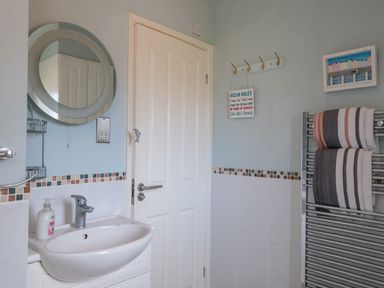 A bathroom with a round mirror above a sink with liquid soap and a door with hooks holding a sign and striped towels on a heated towel rail at High Tide in Rock