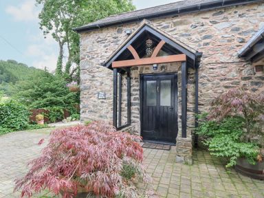 A stone building entrance with a black door and wooden canopy surrounded by plants at The Old Barn in Llanferres near Ruthin