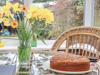 A vase with yellow flowers and a round cake on a glass table with a wicker chair at The Old Barn in Llanferres near Ruthin