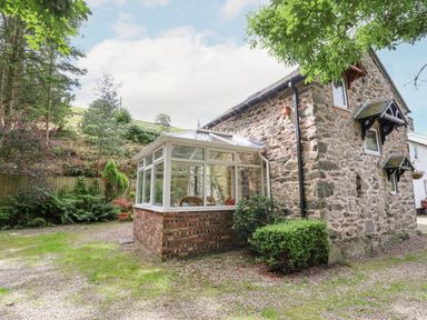 An exterior view of a stone house with a glass conservatory and surrounding garden at The Old Barn in Llanferres near Ruthin