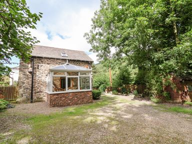 A stone house with a glass conservatory and surrounding garden with trees and shrubs at The Old Barn in Llanferres near Ruthin
