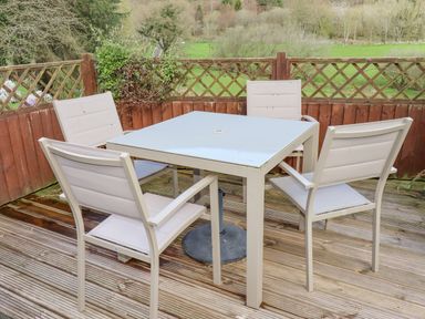 An outdoor patio with four beige chairs around a square glass table on a wooden deck at The Old Barn in Llanferres near Ruthin