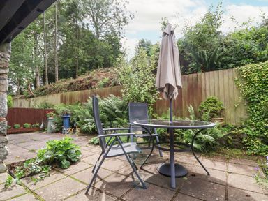 An outdoor patio with a round glass table and two chairs under a closed umbrella surrounded by plants and a wooden fence at The Old Barn in Llanferres near Ruthin