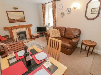 A living room with a dining table set for four a brown leather sofa a fireplace and a small television at Blacksmith Cottage in Appleby-in-Westmorland