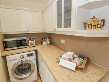 A kitchen corner with white cabinets a washing machine a toaster and microwave on the counter and kitchenware on shelves at Blacksmith Cottage in Appleby-in-Westmorland