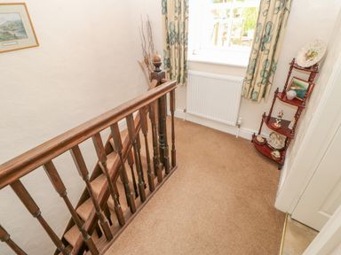 A stair landing with wooden banister a window with patterned curtains a radiator and a corner shelf with decorative items at Blacksmith Cottage in Appleby-in-Westmorland