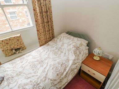 A single bed with patterned bedding next to a wooden nightstand with a decorative lamp by a window with curtains and cushions at Blacksmith Cottage in Appleby-in-Westmorland