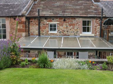 A garden with flowers and grass in front of a stone house with two windows and a transparent roof at Blacksmith Cottage in Appleby-in-Westmorland