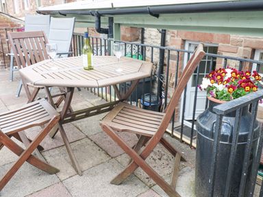 An outdoor patio area with a wooden table and chairs a bottle of wine and two wine glasses and a metal container with flowers at Blacksmith Cottage in Appleby-in-Westmorland