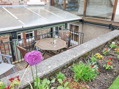 An outdoor seating area with a wooden table and chairs next to a flower bed at Blacksmith Cottage in Appleby-in-Westmorland
